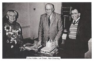 A posed photograph with a cake topped by Standon Church on a table in front of Len Green with Edna Holden on his right and Alan Greening on his left. | H&WLHS